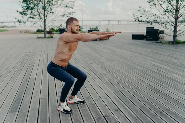 活発な男のフル長射は裸の胴で運動ポーズをしゃがみ込み外に筋肉の身体ポーズを持っています オープンエアのスポーツウェアの演習で強い運動能力の男 自信を持ってフィットネスインストラクター — ストック写真
