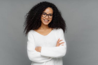 Bukleler ile güzel Afrikalı Amerikalı kadının samimi çekim, nazik bir gülümseme vardır, elleri çapraz tutar, beyaz yumuşak kazak giyer, gri arka plan karşı duruyor. Olumlu duygu ve duygu kavramı