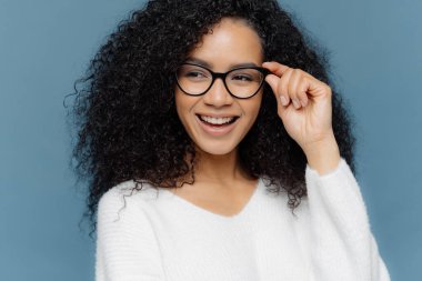 Close up shot of cheerful woman wears glasses, keeps hand on frame, looks gladfully aside, notices something awesome in distance, dressed in casual white jumper, models against blue background