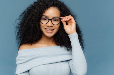 Headshot hoş görünümlü genç Afro-Amerikan kadın gözlük çerçeve üzerinde el tutar, nazik bir gülümseme vardır, mavi kazak giyer, minimal makyaj, sağlıklı bir cilde sahip, mavi arka plan üzerinde izole