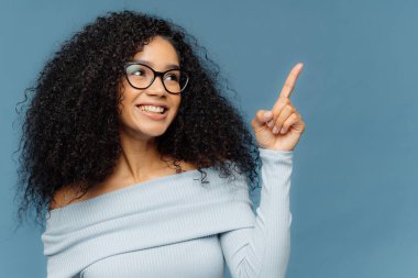 Pleased smiling dark skinned female with Afro hairstyle points index finger upwards, demonstrates something on blank space, has glad expression, wears blue sweater, stands indoor. People and promotion
