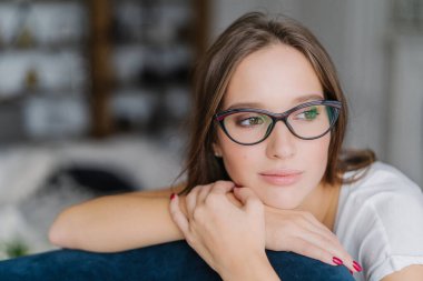 Düşünceli koyu saçlı Avrupa kadın gözlük bir yana görünüyor, kanepenin arkasında, eğilir, gelecekle ilgili contemplates, yalnız hissediyor, yerli atmosfere sahiptir, bulanık arka planı teşkil etmektedir