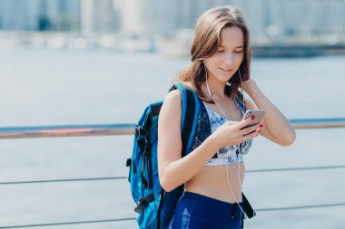 Güzel kadın, mükemmel rakam, yatay portresi tozluk ve üst giyer, göz akıllı telefon odaklanmıştır, büyük sırt çantası taşıyan, kapakları süre doğru yola yalnız uzun mesafe bulmaya çalışır