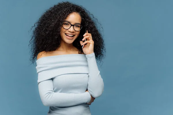 很高兴非洲裔美国女性喜欢电话交谈 保持一只手在腰部 使用现代技术 戴着眼镜和浅蓝色跳线 站在工作室 空白空间为您的口号 — 图库照片