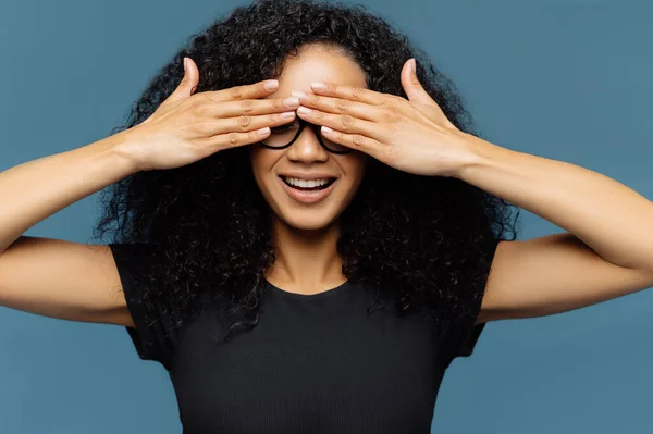 Secret woman with curly hair, covers eyes, wears spectacles, waits for surprise, dressed in black casual t shirt, poses against blue background, has manicure anticipates for miracle. Female hides face