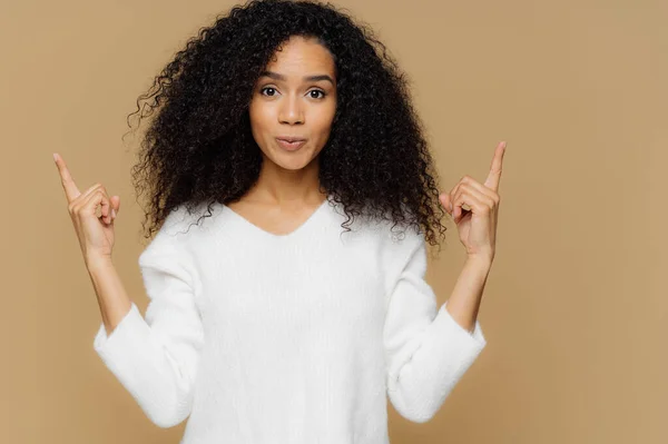照片上自信的女人有浓密的非洲发型 指着两个食指上面 穿着白色跳线 站在棕色背景 广告一些项目 显示方向 上楼来 — 图库照片