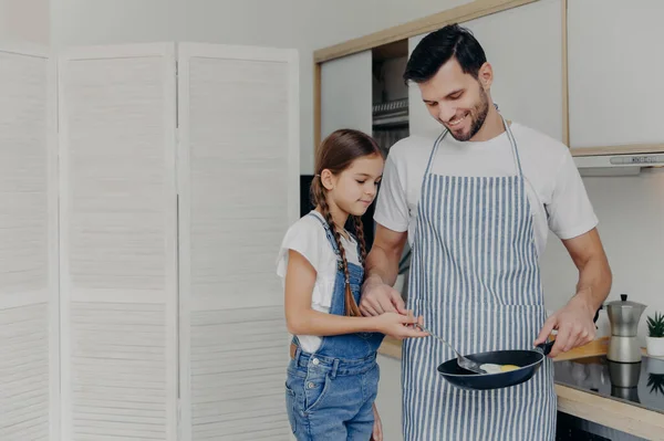 Small female child helps her father to prepare breakfast, fry eggs together, enjoy domestic atmosphhere, stand at kitchen, dad wears apron and holds frying pan. Family and cooking time concept