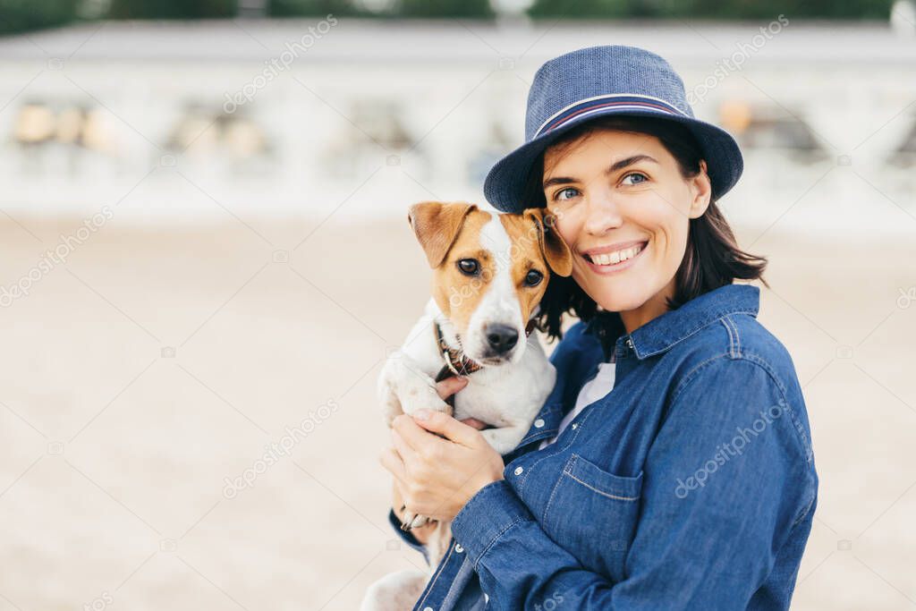 Beautiful young woman has pleasant smile, looks happily aside, holds small dog, enjoys having holidays or weekends, spend time with her favourite pet, poses outdoor. People and animals