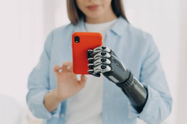 Hands of young european woman with smartphone. Girl is holding the phone with bionic artificial arm and texting. Attractive caucasian woman with artificial limb at home. Modern bionic prosthesis.