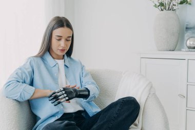 Handicapped girl looks and touches her bionic arm. Young pensive european woman with cyber hand at home. Modern bionic prosthesis. Futuristic technology of artificial limbs.