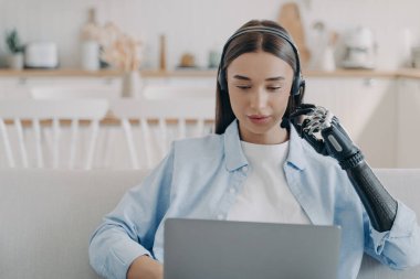 Disabled young woman in headset has online meeting with computer. European handicapped girl has equal rights. Remote manager at home on quarantine. Modern bionic prosthesis technology.