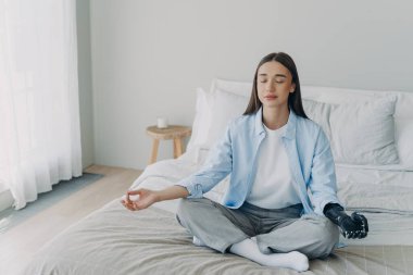 Meditation and concentration. Morning routine of disabled woman. Girl is sitting in lotus pose in her bedroom. Attractive european woman has artificial cyber limb. Stress relief at home.