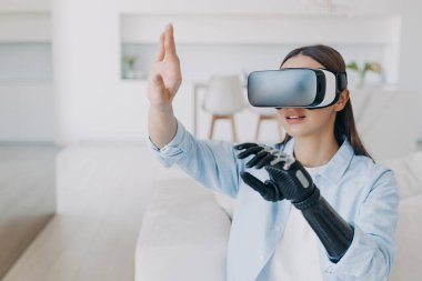 Handicapped girl with cyber bionic hand in virtual reality glasses at home. Disabled person gets rehabilitation. Young european woman sitting in living room on couch. Excited woman touches the vision.