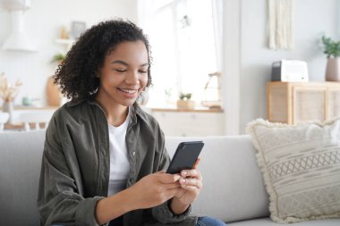 Curly african american girl has fun with phone at home. Happy hispanic woman is browsing social media and smiling. Chatting with friends remotely. Messaging with classmates. Communication concept.