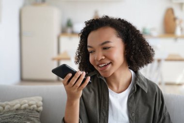 Friendly african american young girl sit on couch at home hold smartphone leave voice message. Modern biracial female chat with friends use loudspeaker virtual assistant search information on internet