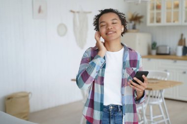 Music listening in airpods on mobile phone. Teenage girl enjoying music in wireless earphones. Afro young woman relaxing at home. Having fun and recreation alone. Music service website or app concept.
