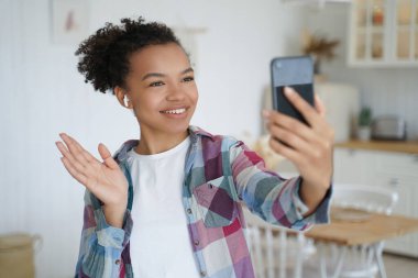 Young mixed race teen girl wear wireless earphones holds smartphone use video call, greet friend, waving hand. Friendly biracial female blogger start talk in social network, welcoming followers.