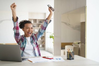 Excited happy african american girl student got good exam test scores on laptop. Overjoyed teenage schoolgirl raising hands screaming celebrates personal achievement or finishing homework.