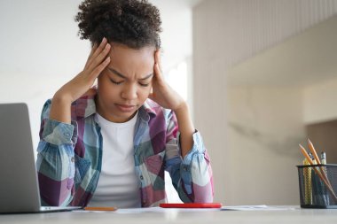 Tired african american teenage schoolgirl suffer headache while doing homework at laptop at home. Exhausted, frustrated teenager girl worried about educational problem or bullying. Difficult elearning