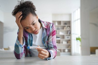Anxious teenage girl gets positive pregnancy test result. Desperate african american young woman is holding stripe test and looking at it. Teenage lifestyle and mistakes. Contraception concept.