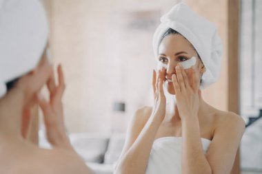 Girl applies eye patches and looking into mirror. Attractive european woman wrapped in towel after bathing. Young hispanic lady takes shower at home. Relaxation at spa resort. Beauty routine at home.