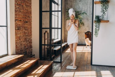 Anxious slim caucasian woman taking step onto scale to measure her weight. Girl wrapped in towel after bathing has time at spa. Young woman checking weight after shower. Fitness and wellness concept.