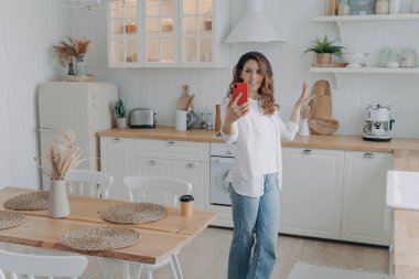 Young hispanic woman boasting with her stylish kitchen. Housewife has video phone call at home. Girl shows modern white scandinavian interior into camera. Stove, worktop and cuisine.