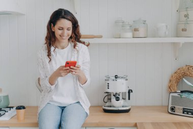 Attractive young woman is texting on smartphone. Housewife has leisure and sitting on worktop at kitchen at home. Smiling girl with telephone. Modern luxurious scandinavian interior.