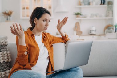 Puzzled young woman has online conference at home. European lady with laptop is talking at internet meeting. Confused girl is telling a problem. Businesswoman is working while quarantine.