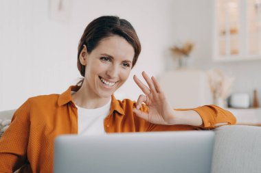 Young woman has video call and shows ok. European girl has online conference at home. Freelancer or businesswoman with computer is talking at internet meeting. Remote work on quarantine.