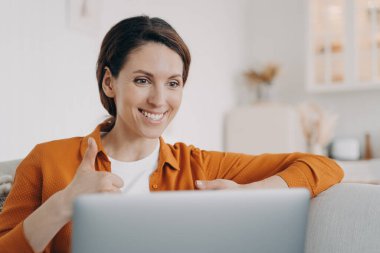 Young spanish woman is speaking online and showing thumb. Girl has online conference indoors. Lady with laptop is talking at internet meeting. Working on a couch at home while quarantine.