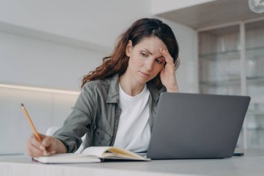 Overworked spanish lady has got a headache. Sick tired freelancer is working at home. Exhausted woman is chatting on laptop and taking notes with pencil. Frustrated businesswoman has problem.
