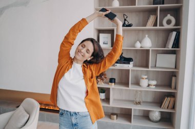 Happy european woman is dancing alone at home. Girl in earphones is having fun in her living room. Music listening online. Concept of weekend, relaxation and emotion expression. Cosy home interior.