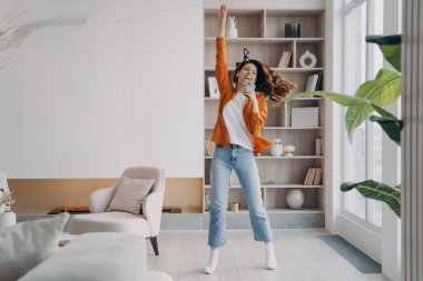Young woman is singing, dancing and jumping. Carefree girl with smartphone having fun crazy and screaming at home. Favourite song and relaxation. Concept of emotional expressions and rebellion.