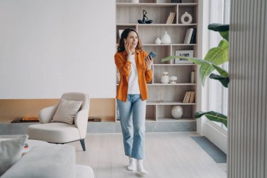 Happy european girl has phone call at home. Pretty young woman is using airpods. Wireless earphones. Technology using, leisure and communication concept. Beautiful modern interior.