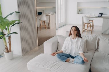 Young hispanic woman is practicing yoga and meditation at home. Girl in white is sitting on couch with her eyes closed. Posture exercise, lotus asana, gymnastics. Wellbeing and morning routine.