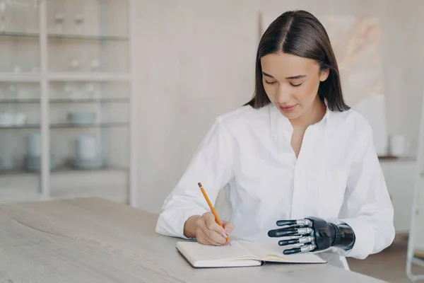 Disabled european girl is taking notes at the desk and doing homework. Happy handicapped young woman with futuristic cyber arm is studying remotely in college. Concept of equal opportunities.