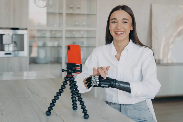 Disabled european woman is streaming video in social media and laughing. Attractive handicapped girl is taking video how to use cyber arm. Beauty blogger has modern myoelectric prosthesis.