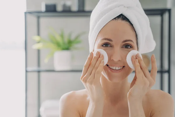 Girl is cleansing skin with face toner and cotton disks. Attractive caucasian woman wrapped in towel after bathing. Young woman takes shower at home. Delicate skin care and rejuvenation.