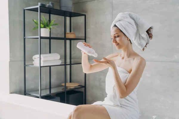 Body lotion applying from bottle. Attractive caucasian girl wrapped in towel after bathing. Happy young woman takes shower at home. Products for smooth silky skin using. Spa procedure.