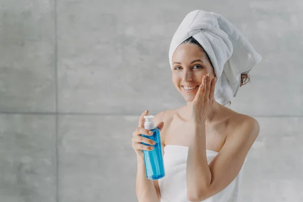 Face toner or micellar water advertising concept. Happy young woman touches pure skin. Gorgeous caucasian woman wrapped in towel after bathing applying toner in flacon. Face washing foam in bottle.