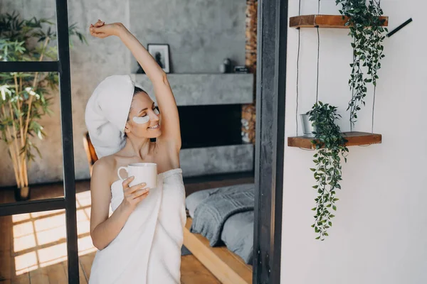 Young woman with cup of tea relaxing. Vacation at spa resort. Girl applies anti wrinkle eye patches. Attractive european woman wrapped in towel after bathing enjoying pure skin and beauty procedures.