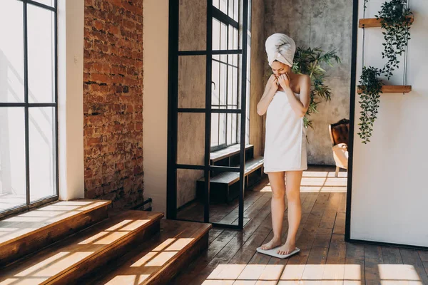 Weight gaining concept. Frustrated slim european woman taking step onto scale to measure her weight. Girl wrapped in towel after bathing has time at spa. Stressed woman checking weight after shower.