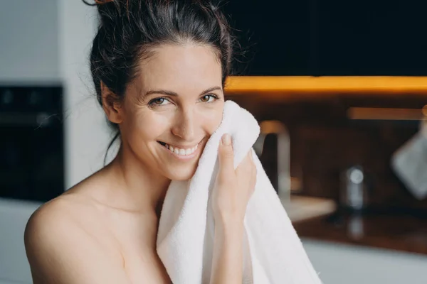 Caucasian woman is wiping face with towel after washing and smiling. Young woman takes shower at home and doing daily skin care. Hygiene and wellness, dermatology and skin cleansing.