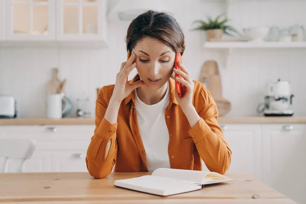 Tired young woman is remote employee. Girl is working from home sitting at the table and talking on telephone. Secretary, office manager or customer support assistant. Workspace at home concept.