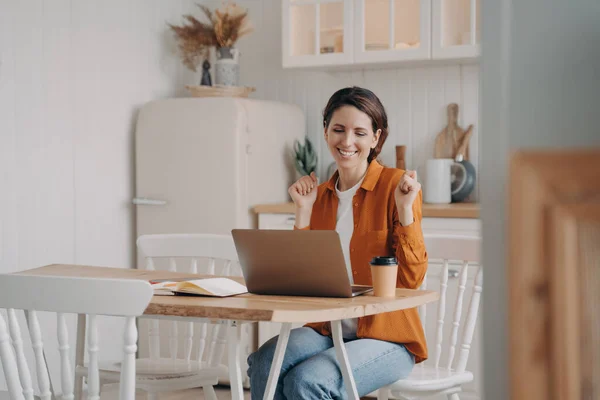 Business lady has online conference. Confident young woman wins. Girl is working in front of computer. European woman is happy. Distance work at the kitchen on quarantine concept.