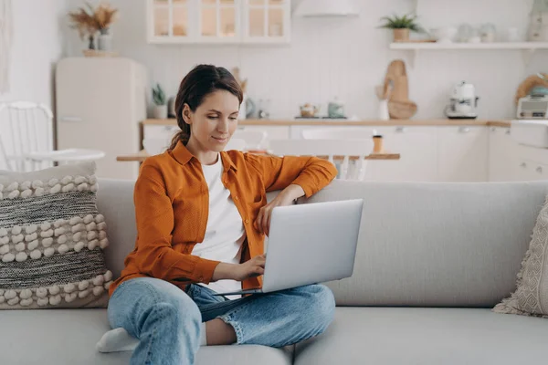 Happy young woman with laptop is sitting on couch at home. Spanish girl is chatting and browsing internet. Remote work or study from home. Cosy living room and kitchen interior.