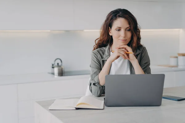 European girl is sitting in front of screen with hands folded. Confident female executive is working with laptop at home. Successful lady boss at her kitchen. Remote work of lawyer assistant.