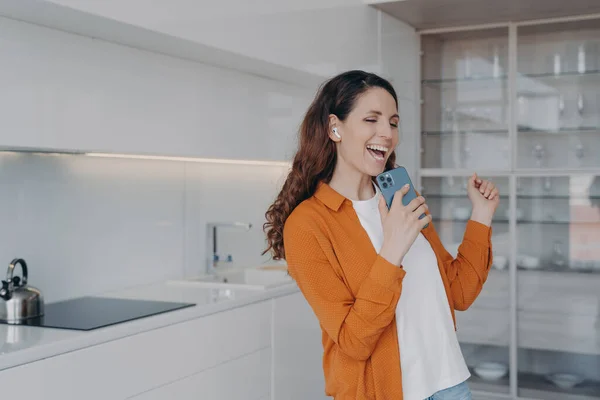 Happy european woman is singing with phone as with microphone. Girl having fun in her living room. Application for music listening online. Favourite song singing at home, relaxation and dancing.