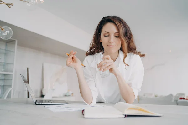 Young spanish woman in white is taking notes. Teacher is checking schedule. Girl is writing down into notebook at the desk preparing to online lesson. Remote work and study from home on quarantine.